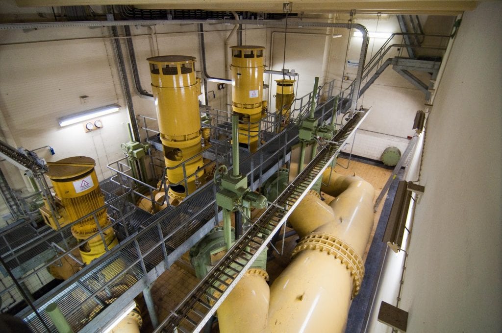 Overhead view of a heavy-industrial pumping-station interior with large yellow vertical pumps and pressurised pipework.